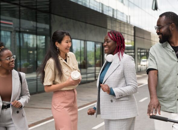 Four diverse professionals are walking and having a conversation outside of a modern glass building. They are smiling and appear to be in a lively discussion.