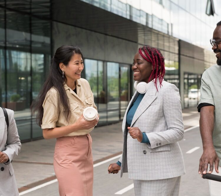 Four diverse professionals are walking and having a conversation outside of a modern glass building. They are smiling and appear to be in a lively discussion.