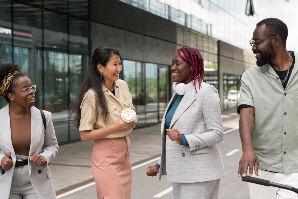 Four diverse professionals are walking and having a conversation outside of a modern glass building. They are smiling and appear to be in a lively discussion.