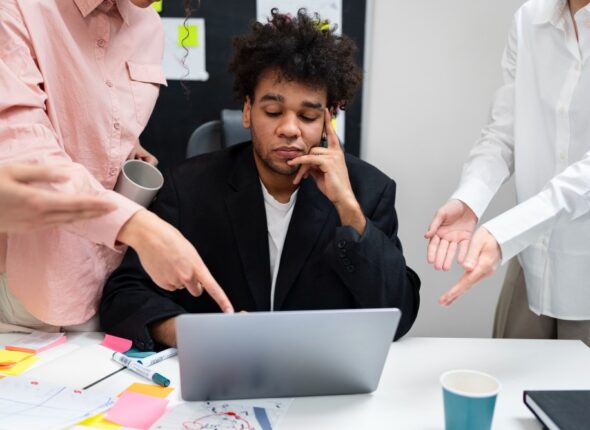 A young man looking tired or frustrated at his laptop while surrounded by colleagues gesturing and discussing work in a busy office environment.