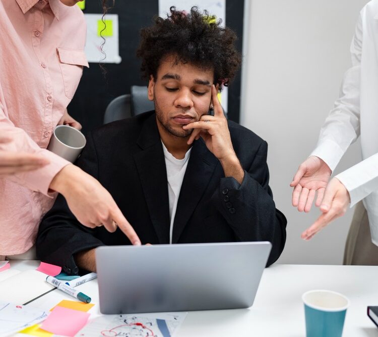 A young man looking tired or frustrated at his laptop while surrounded by colleagues gesturing and discussing work in a busy office environment.