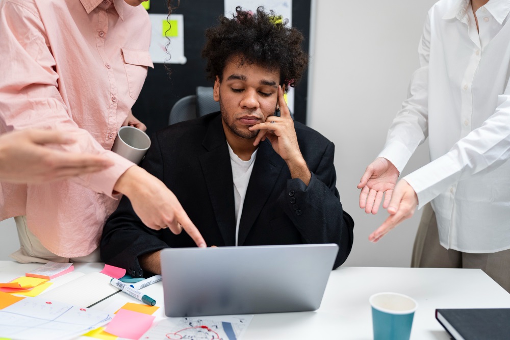 A young man looking tired or frustrated at his laptop while surrounded by colleagues gesturing and discussing work in a busy office environment.