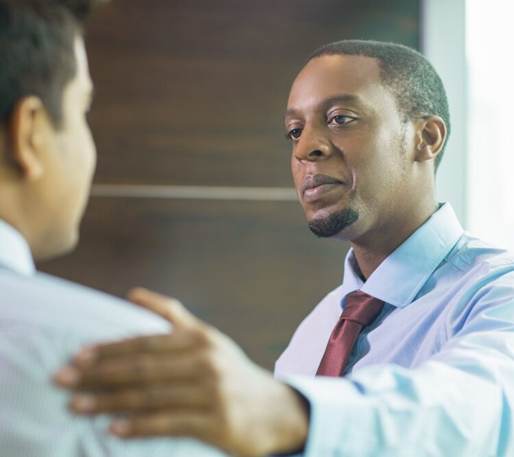 Two men in business attire having a conversation, with one man gesturing towards the other's shoulder in a supportive or guiding manner.