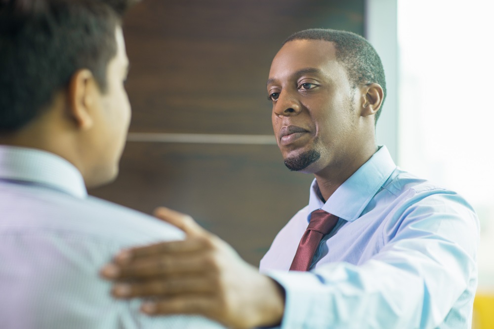 Two men in business attire having a conversation, with one man gesturing towards the other's shoulder in a supportive or guiding manner.