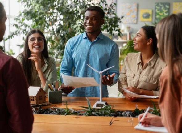 A group of diverse professionals in a friendly discussion around a table, with some holding papers and smiling, amidst model houses and office supplies, in a well-lit room with plants and art on the walls.