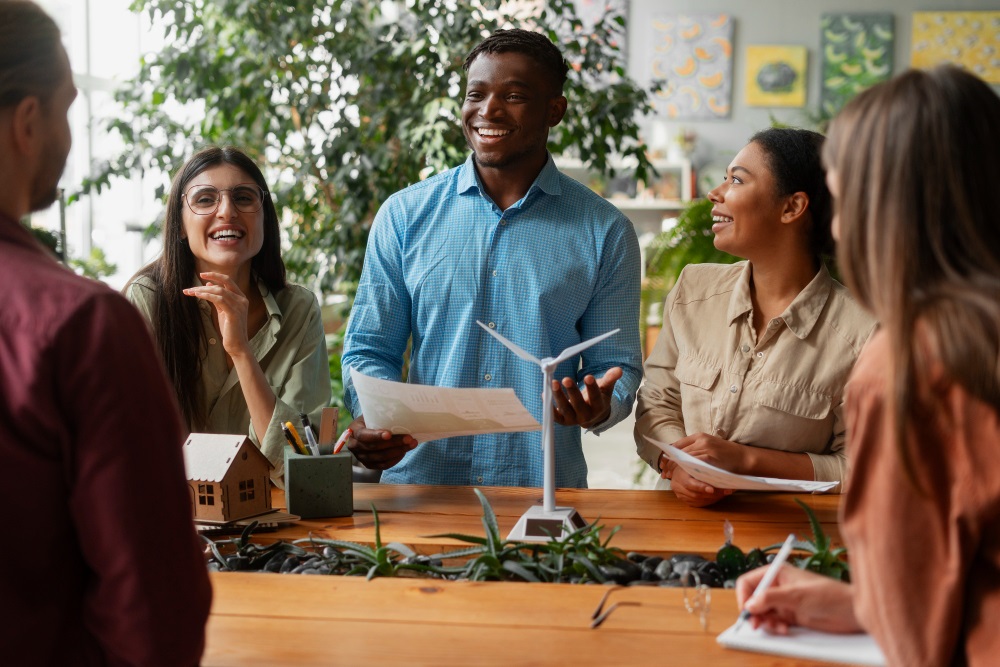 A group of diverse professionals in a friendly discussion around a table, with some holding papers and smiling, amidst model houses and office supplies, in a well-lit room with plants and art on the walls.