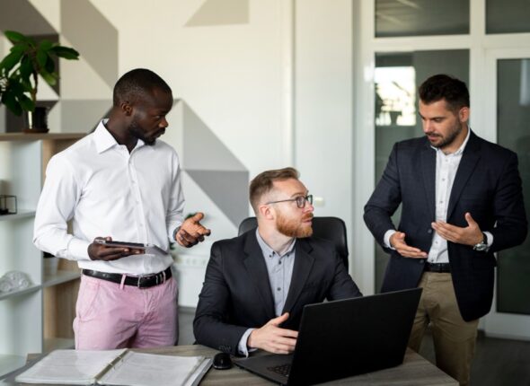 Three men in professional attire stand around a laptop on a desk in a modern office, engaged in a discussion.