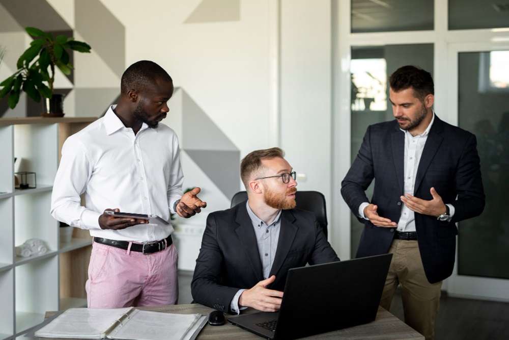 Three men in professional attire stand around a laptop on a desk in a modern office, engaged in a discussion.