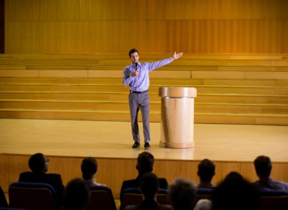 A man presenting on stage to an audience with one arm extended, standing near a wooden podium in a lecture hall.