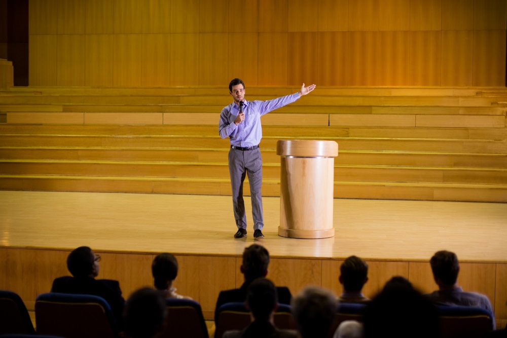 A man presenting on stage to an audience with one arm extended, standing near a wooden podium in a lecture hall.